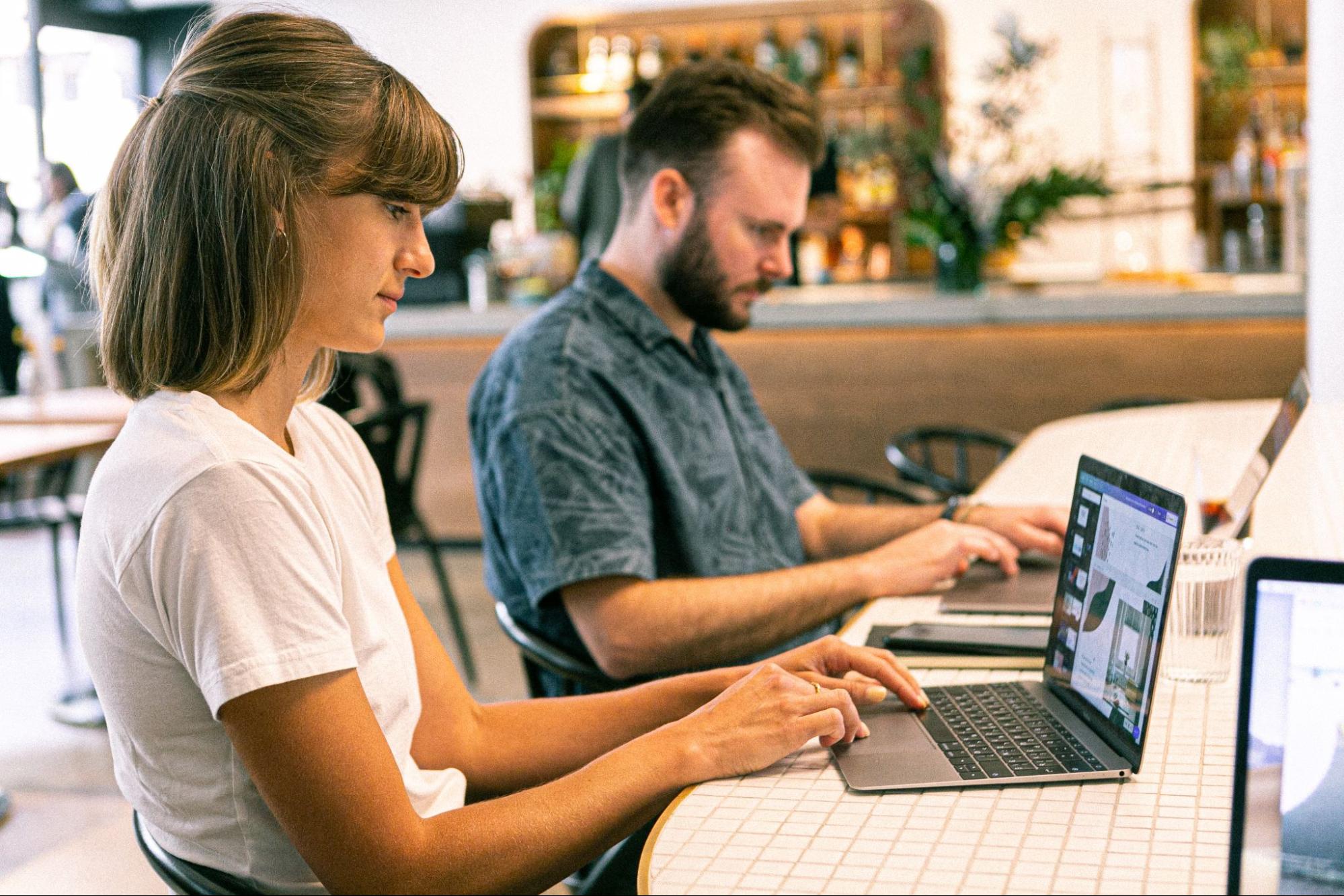 Homem e mulher utilizando computadores portáteis enquanto estão sentados em uma mesa.