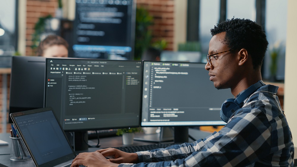 Imagem de um homem negro engenheiro de software trabalhando com 3 telas em um computador