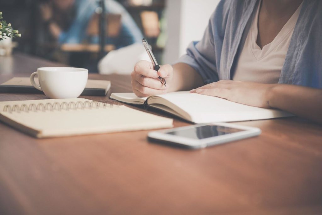Mão humana feminina com uma caneta na mão em uma mesa de estudos com cadernos, celular e uma xícara de café