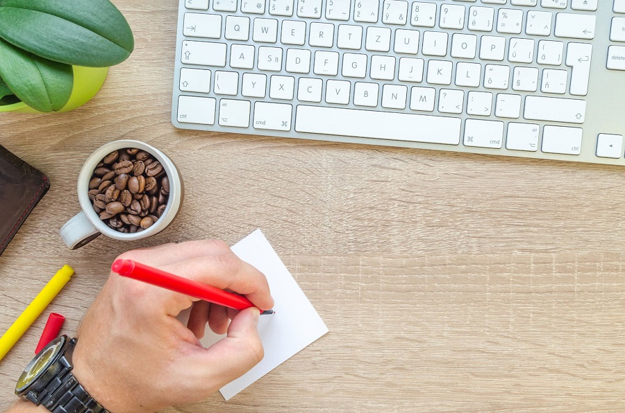 Homem escreve em post-it. Na foto, há um teclado e uma xícara com grãos de café.