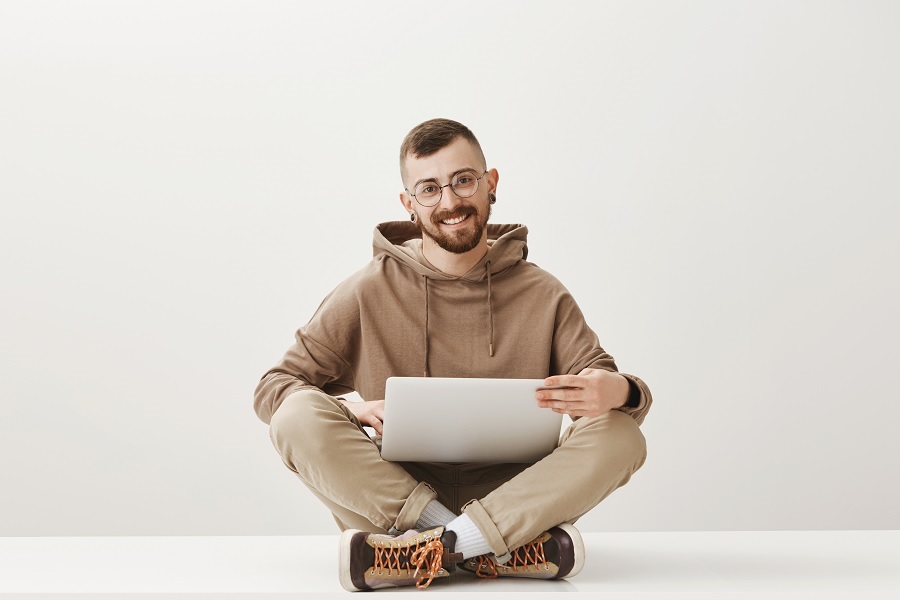 Foto de um rapaz branco de óculos sorrindo sentado de pernas cruzadas com um notebook no colo.