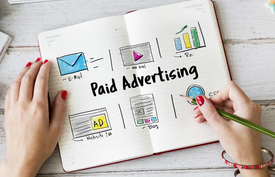 Mulher escreve em caderneta o termo "Paid Advertising", com alguns ícones ao redor indicando a técnica de mídias pagas.