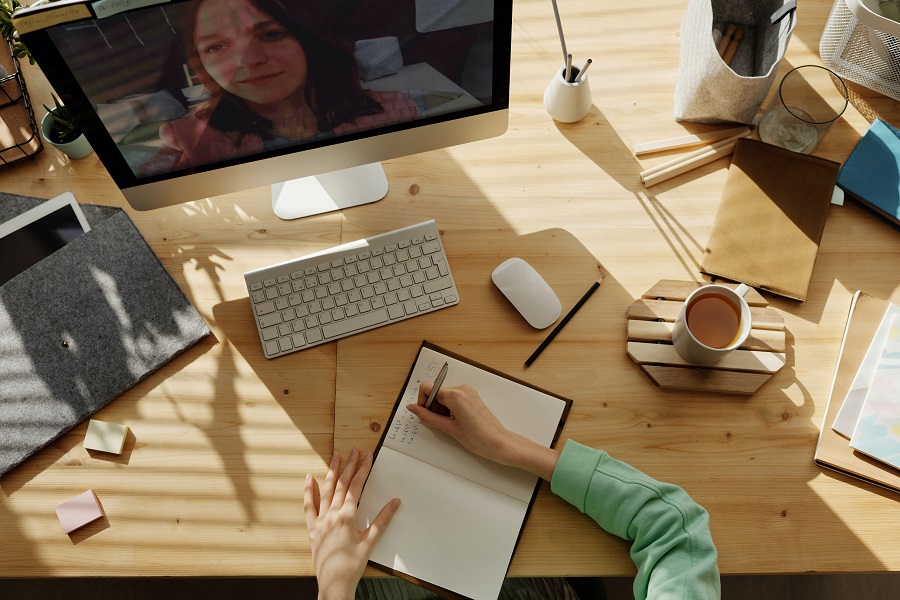 Pessoa escrevendo em caderno enquanto assiste um vídeo, ou conversa com alguém, via computador.