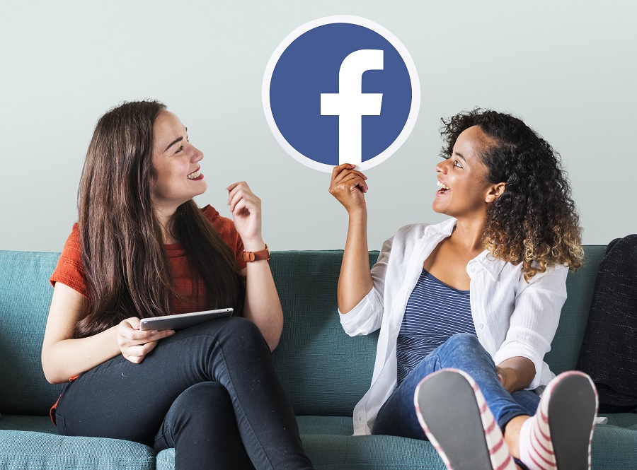 Duas mulheres sentadas em um sofá sorrindo e segurando o símbolo do Facebook.