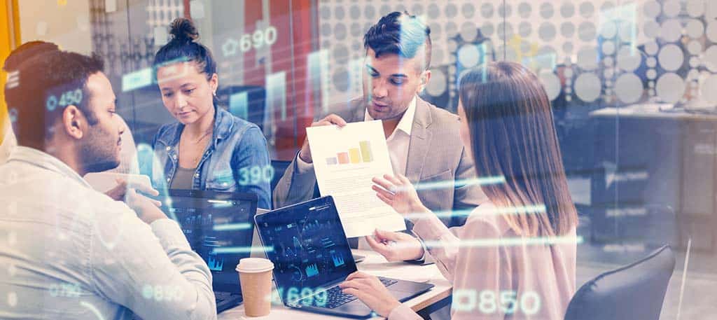 Equipe discutindo tecnologia em escritório com gráficos digitais sobrepostos, representando o que é virtualização de hardware e a otimização de recursos computacionais.