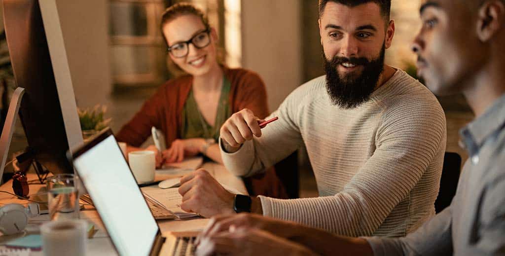 Equipe de três profissionais jovens colaborando em um projeto de arquitetura e desenvolvimento de software enquanto discutem estratégias para otimizar sistemas, representando a importância dessa área no mercado atual.