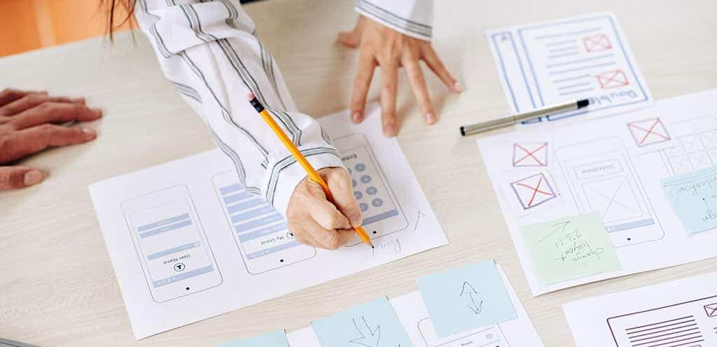 Desenvolvedor de apps enquanto trabalha e esboça o design e a interface de um aplicativo móvel em papéis sobre a mesa.
