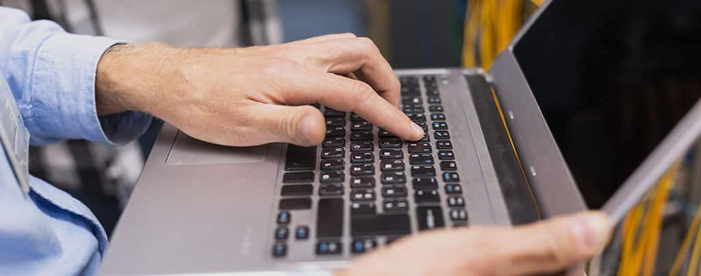 Profissional em redes de computadores configurando um sistema em um laptop, com servidores e cabos ao fundo.