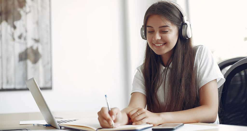Jovem mulher sentada à mesa fazendo anotações em um caderno enquanto estuda arquitetura de software em seu laptop.