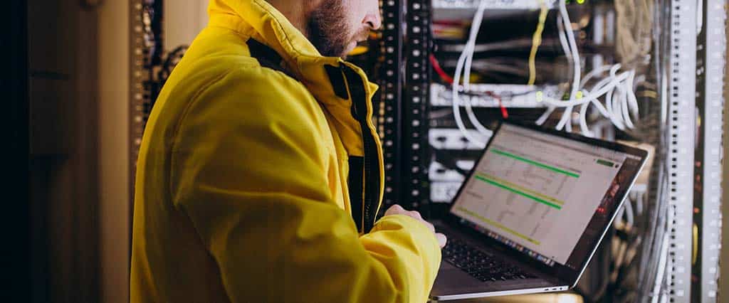 Engenheiro de TI analisando dados em um laptop dentro de um data center, ilustrando como funciona a virtualização de servidores e o gerenciamento remoto.