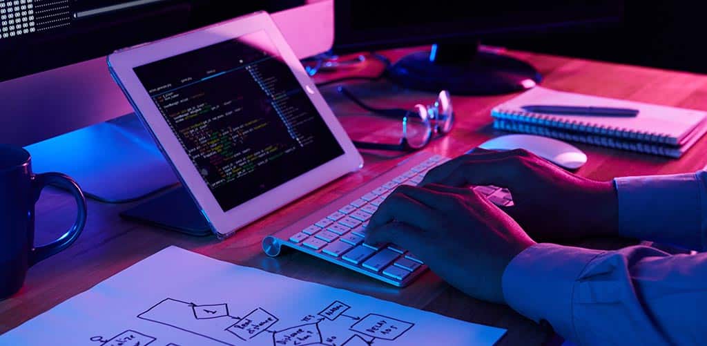 Desenvolvedor programando em um tablet com fluxograma ao lado, representando a adoção de uma visão de implementação em arquitetura de software para organização de um sistema.