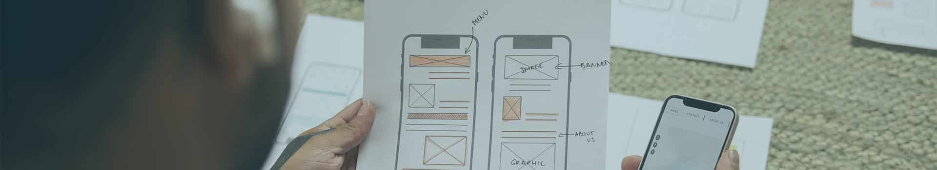 Pessoa analisando um esboço físico de interface enquanto testa um aplicativo no celular, demonstrando o processo de criar app.