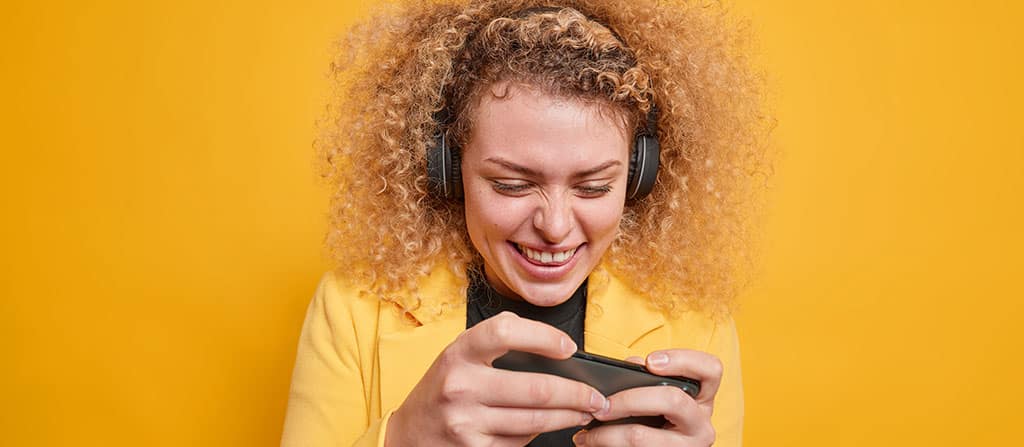 Mulher sorrindo enquanto joga no celular, representando o resultado do trabalho de quem aprende como criar um app de jogo.
