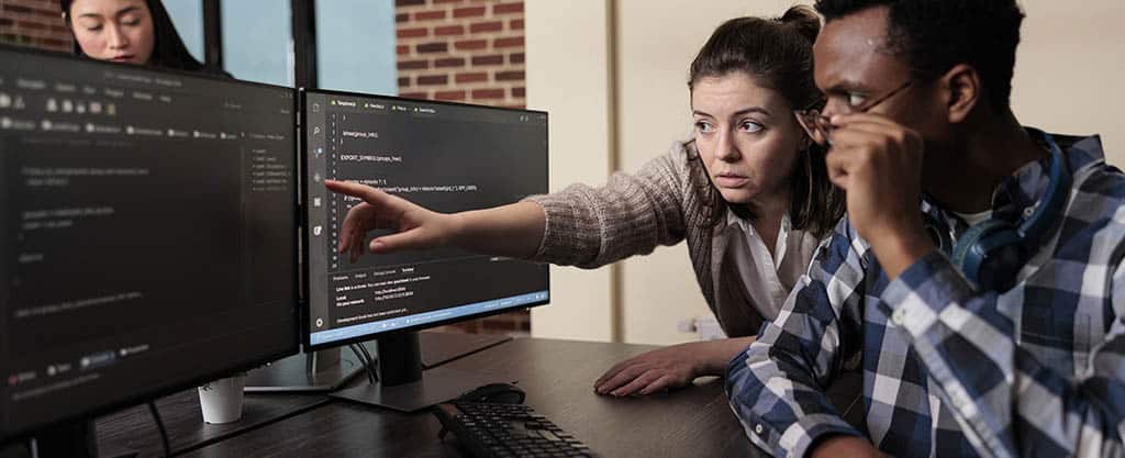 Equipe de tecnologia analisando código em monitores, representando o dia a dia de trabalho colaborativo de um programador back end no desenvolvimento de sistemas.