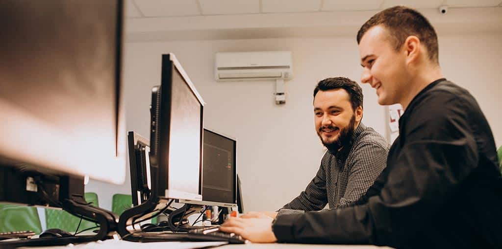 Dois desenvolvedores colaborando em engenharia de software e projeto de arquitetura, analisando e discutindo soluções em frente a monitores no escritório.