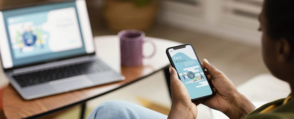 Pessoa sentada usando um smartphone para aprender como registrar um aplicativo, com um café e um laptop aberto ao fundo para ajudar no processo.